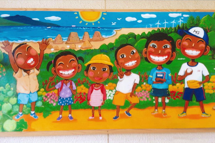 三種町・浜口小学校に展示されたアクリルイラスト。釜谷浜をイメージした、サンドクラフトと風力発電が描かれた海岸線と、緑豊かで花溢れる大地を背景に、日焼け肌の六人の小学生たちが、RiRiKOの持ち味であるコミカルな笑顔で描き出されている。