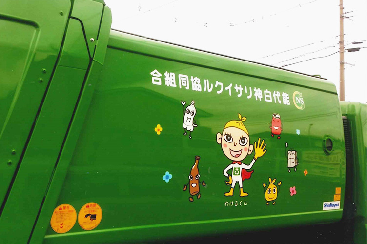 能代市リサイクルセンター（能代白神リサイクル組合）で利用されている、ゴミ収集車の写真。緑色の車体の側面には、びん、カン、ペットボトルなどのリサイクルごみ達をモチーフにした可愛らしいキャラクターたちと、イメージキャラクター「わけるくん」が描かれている。