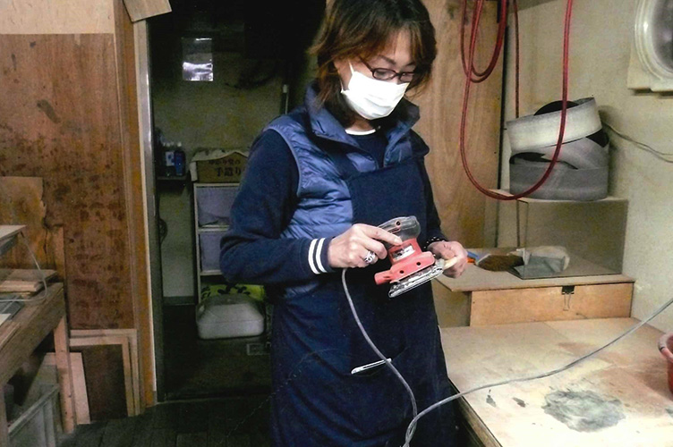 木工作業場で秋田杉の木片を磨き上げるRiRiKOをやや引きで撮影したウエストショット写真